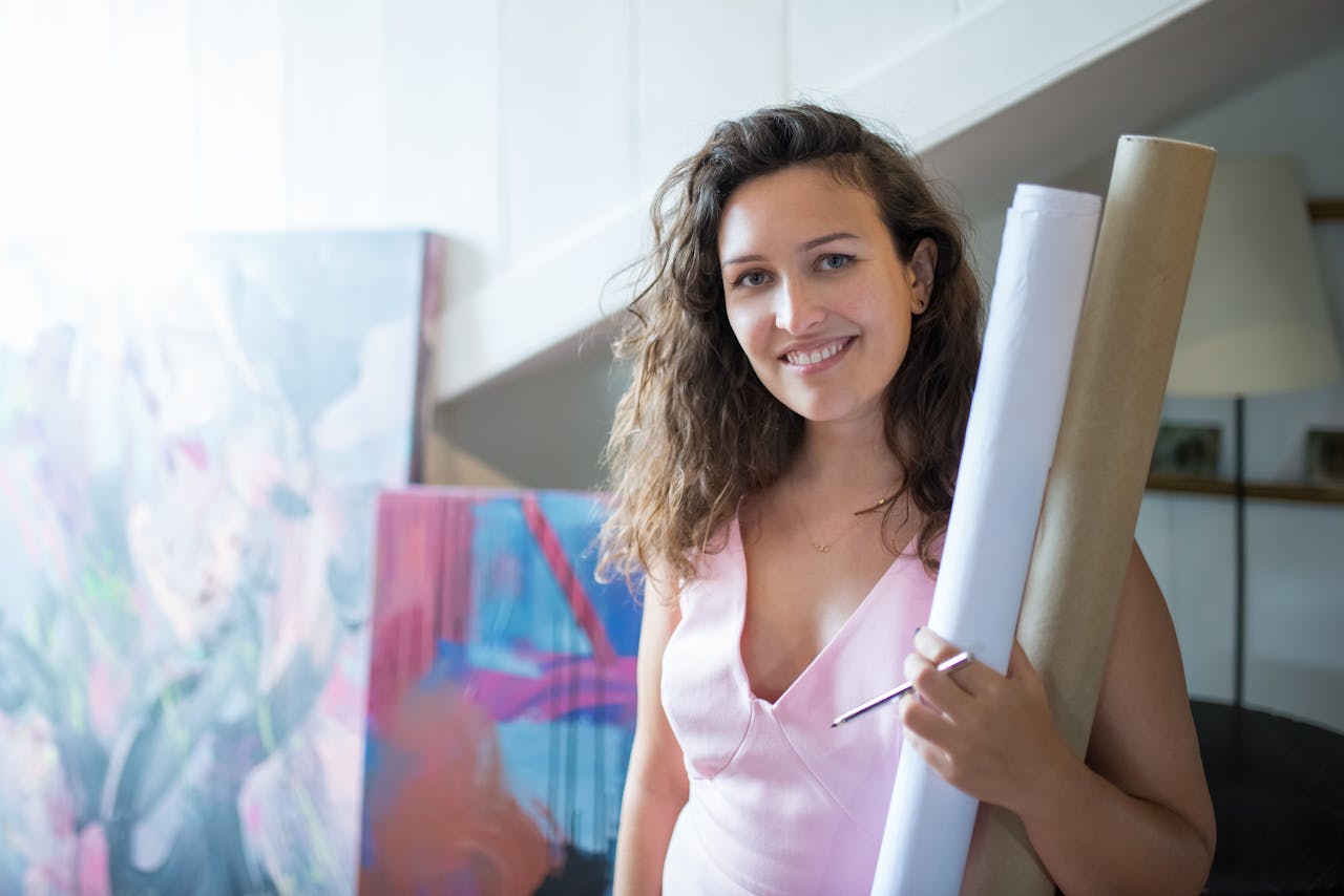 Smiling woman artist in studio holding canvas and paper rolls, exuding creativity and professionalism.